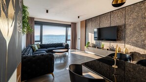 A modern living room with a large window, a black sofa, a dining area, and a TV mounted on the wall.