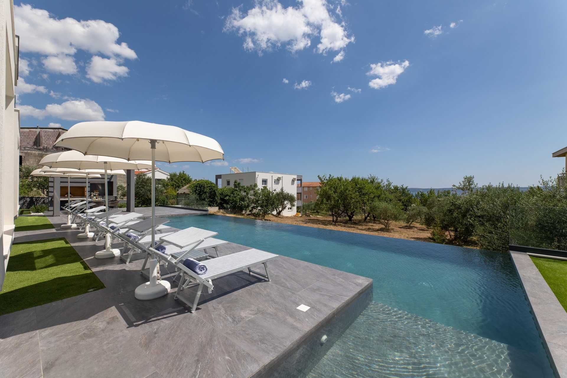 Uma área de piscina com espreguiçadeiras e guarda-sóis, cercada por vegetação e edifícios.
