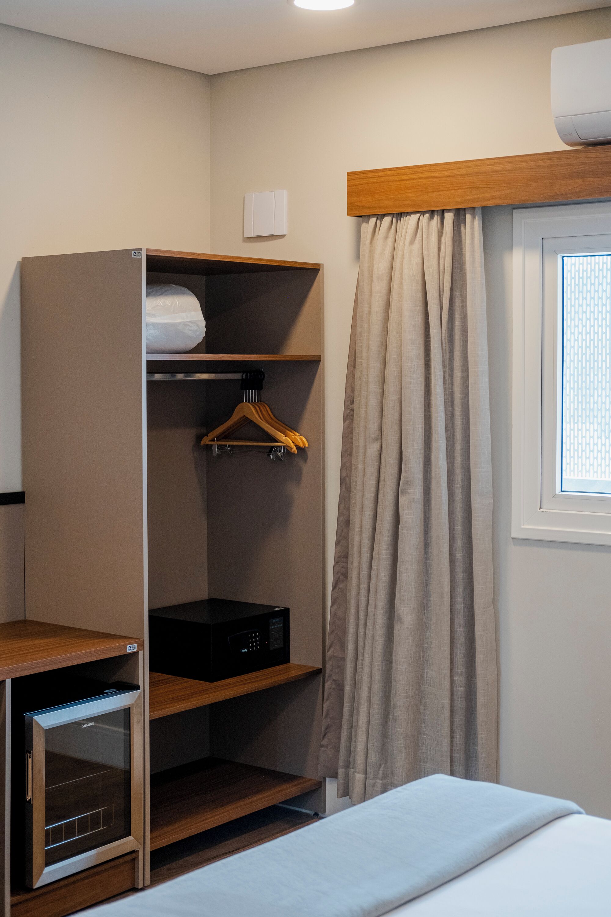 A compact room with a bed, a built-in microwave, a wooden wardrobe, and a window with curtains.
