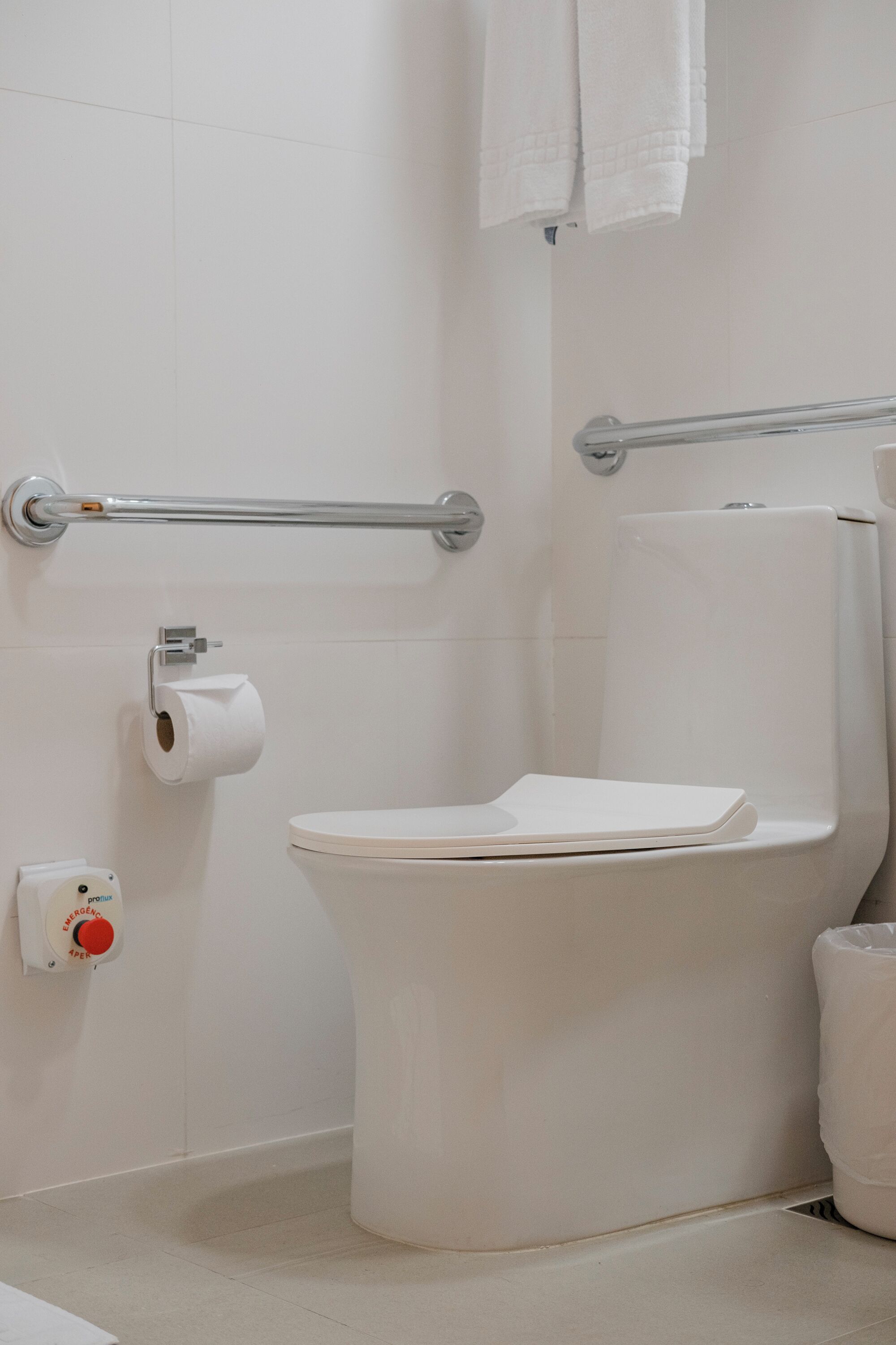 A bathroom with a white toilet, a handrail, a roll of toilet paper, and an emergency button.