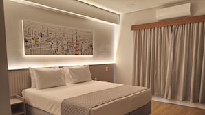 A modern hotel room with a large bed, a headboard with built-in lighting, a wall-mounted air conditioner, and a cityscape artwork above the bed.