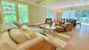 A spacious living room with a beige sofa, two armchairs, a coffee table, and a dining area with a table and chairs. Large windows provide natural light and a view of greenery outside.