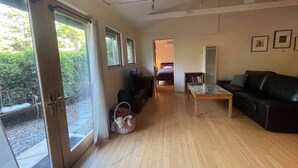 A spacious living room with a large sliding glass door leading to a balcony, a black leather sofa, a small table, and a bed in the background. - Charming guest cottage in Santa Barbara with access to hiking and the  beach (Santa Barbara)