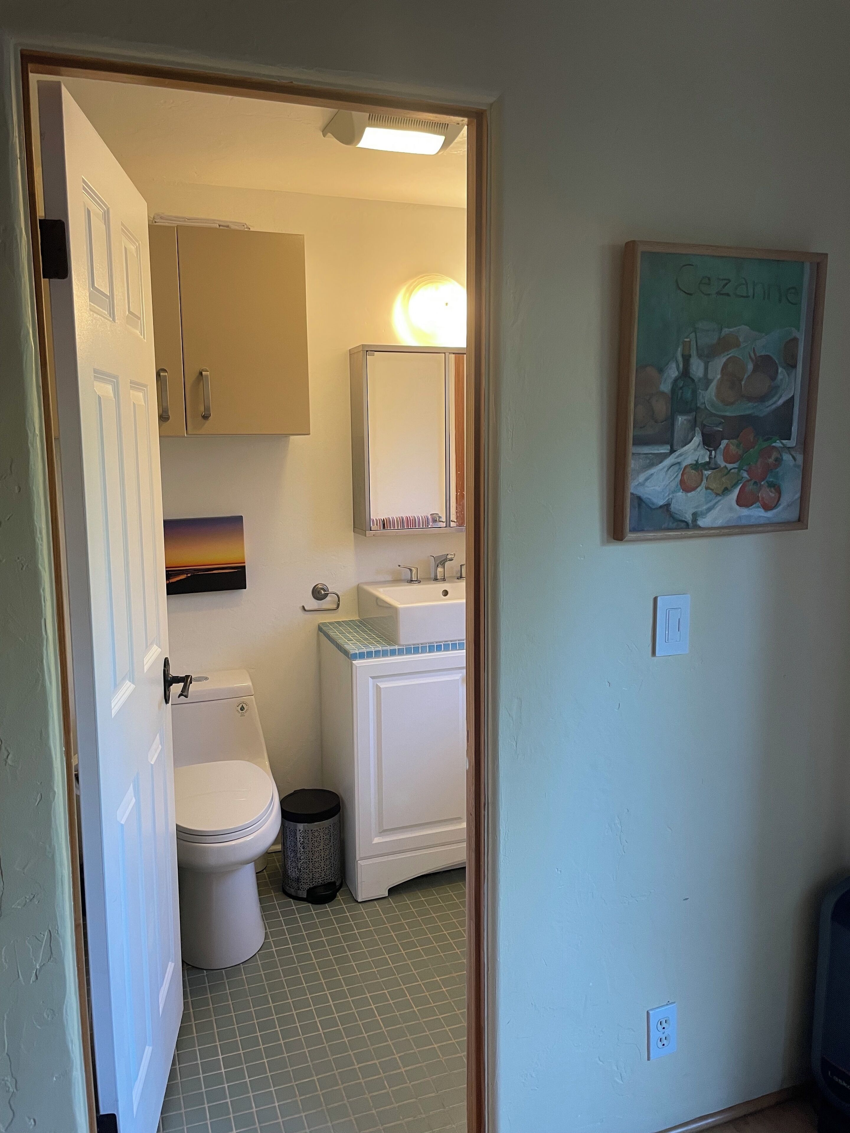 Une salle de bain avec des toilettes, un lavabo et un miroir. On y trouve également une poubelle et un tableau accroché au mur.