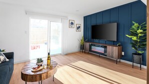 A modern living room with a wooden floor, a blue accent wall, a flat-screen TV, a wooden coffee table with a bottle and glasses, and a sofa. - MCM Condo Near DT SoCo Pet Friendly (Austin)