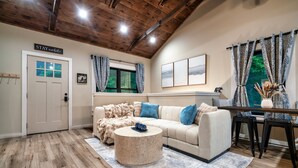 A cozy living room with a wooden ceiling, a beige sofa with blue cushions, a round coffee table, and a view of greenery through the window. - Modern Cabin Near Pigeon Forge, Sleeps 8, Hot Tub, Game Room & Fire Pit (Sevierville)