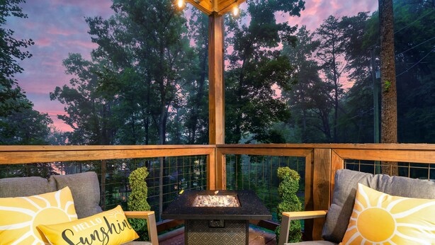 An outdoor seating area with two cushioned chairs and a small table, surrounded by a wooden deck and glass railings, overlooking a forested area. - Modern Cabin Near Pigeon Forge, Sleeps 8, Hot Tub, Game Room & Fire Pit (Sevierville)