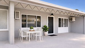 Una casa con un porche techado, una mesa de comedor con sillas, una planta en maceta y un equipo de aire acondicionado.