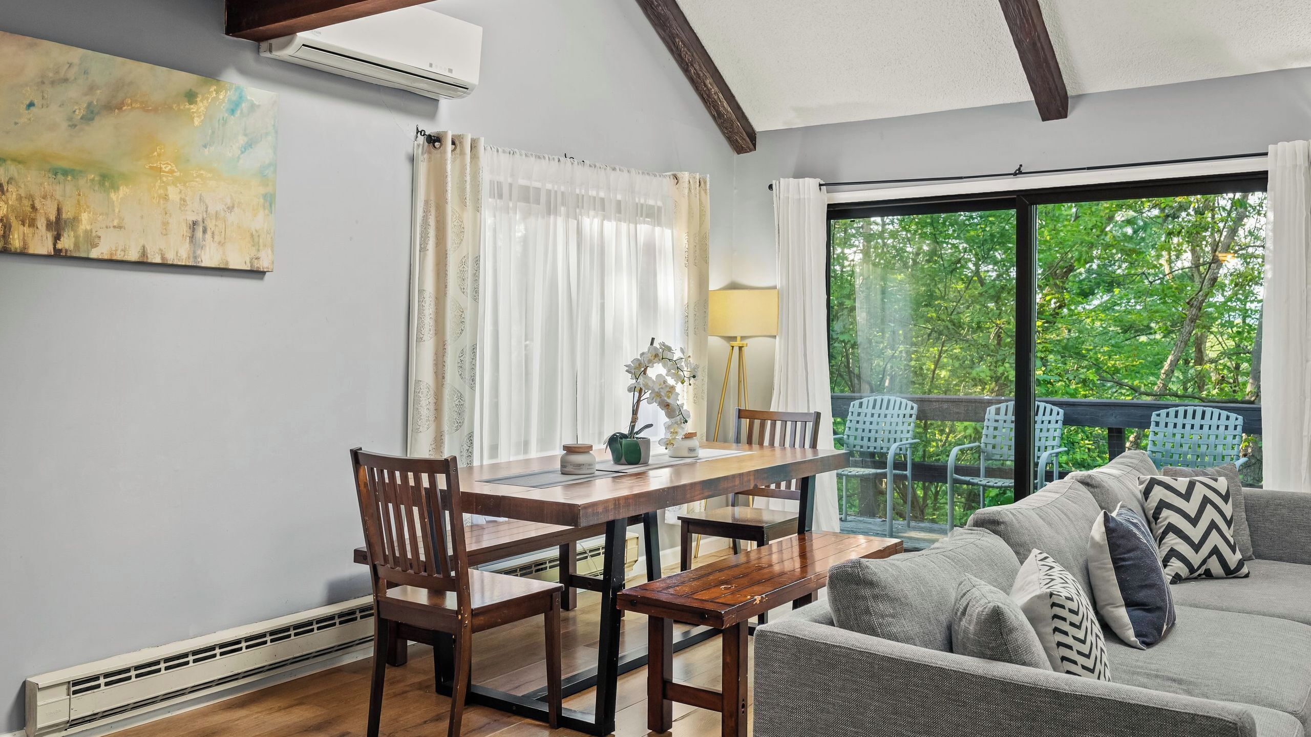 Un coin repas avec une table et des chaises en bois, un canapé agrémenté de coussins à motifs, une lampe et une vue sur la verdure grâce à une grande fenêtre.