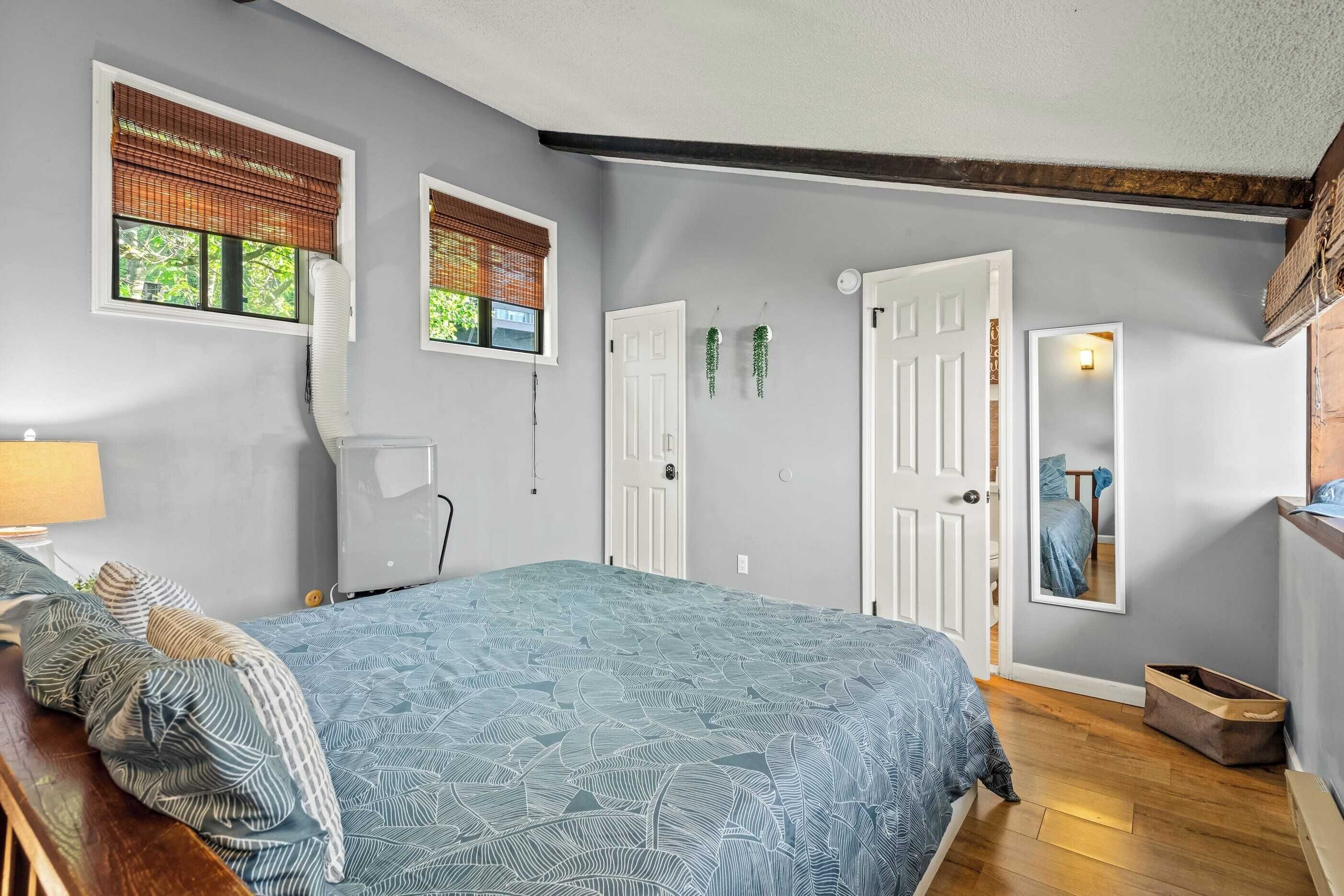 A bedroom with a bed, a standing fan, two windows with blinds, a door, and a mirror.
