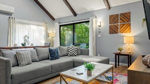 Un salon moderne avec un canapé d’angle gris, une table basse en marbre et une vue sur la verdure à l’extérieur.