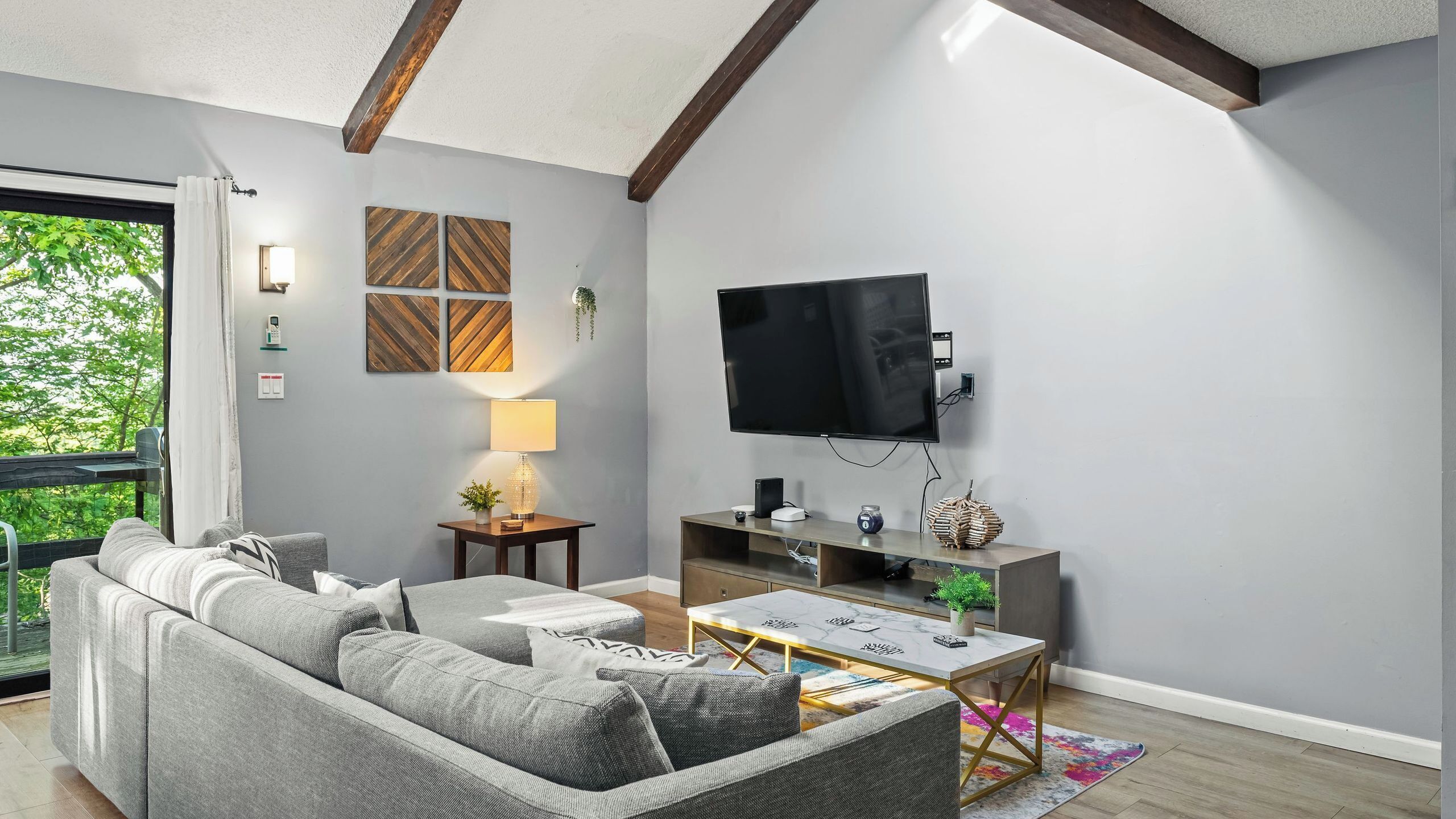 A modern living room with a grey sofa, a flat-screen TV, a small table, and a window with curtains.