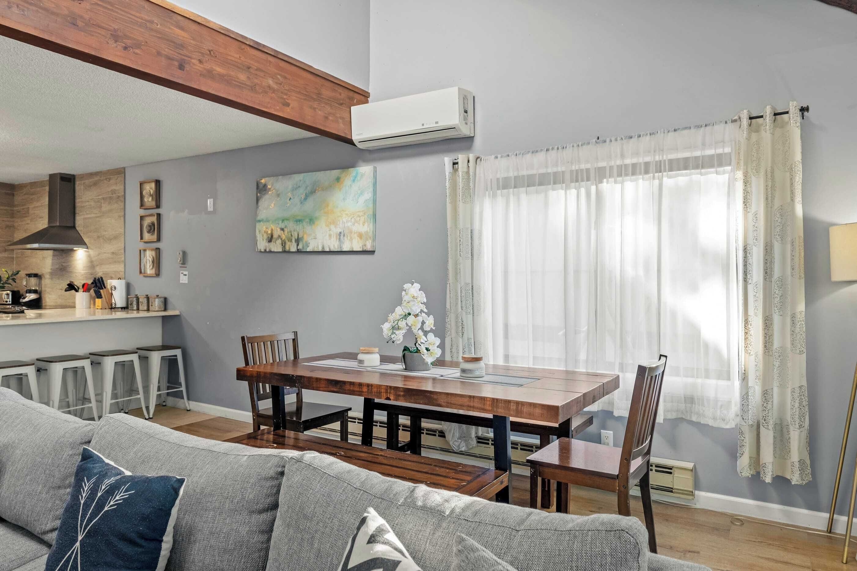 A modern living room with a wooden dining table, grey sofa, and a kitchen area with bar stools.