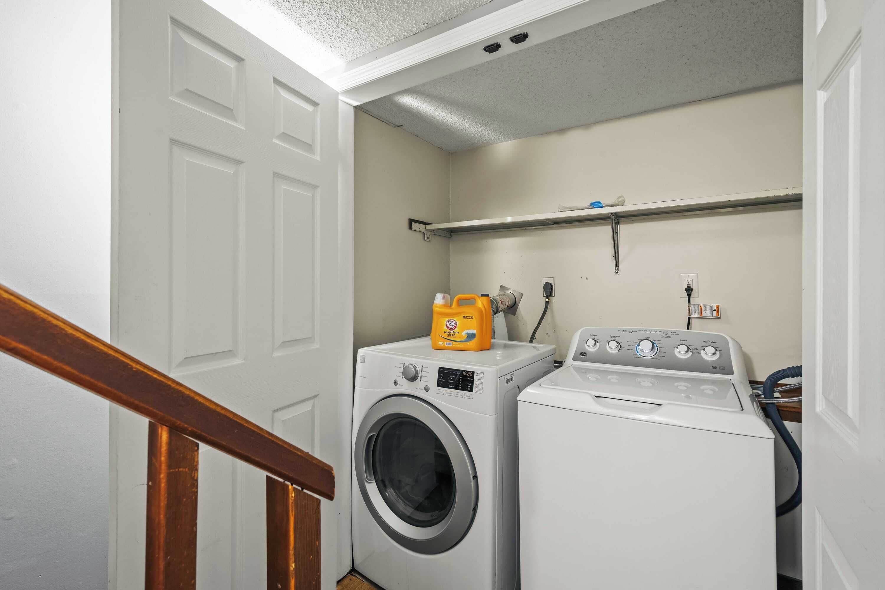 A laundry room with a front-loading washer and dryer, a shelf above the dryer, and a wooden staircase leading to another room.
