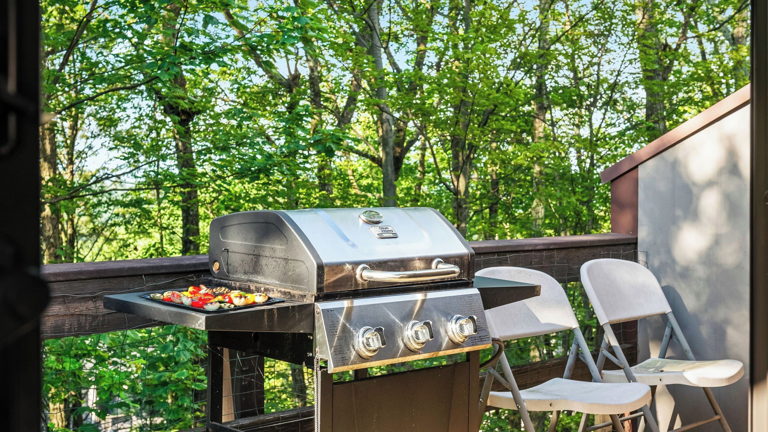 An outdoor grill with a view of trees, a chair, and a wooden deck.