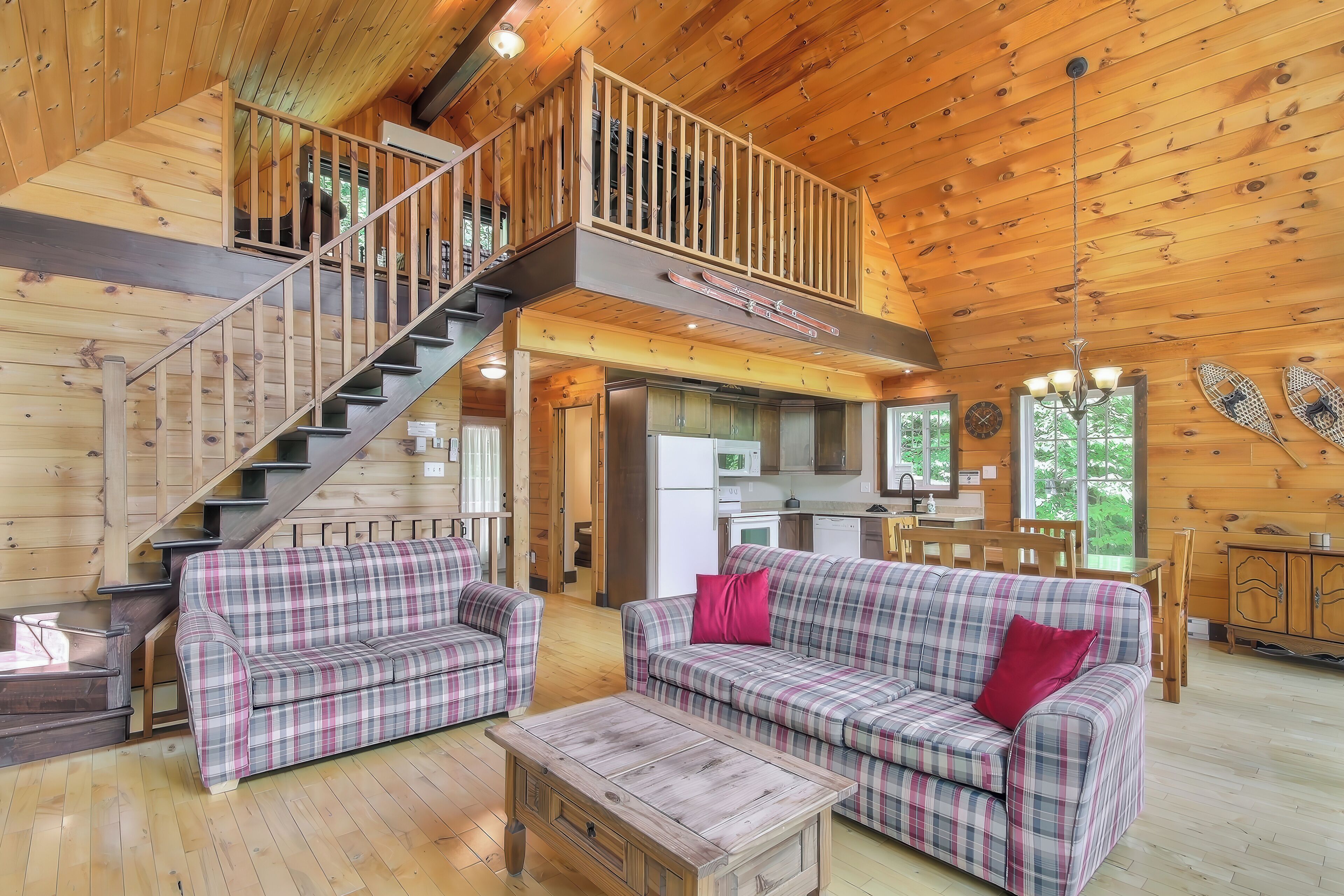 A wooden cabin interior with a staircase, two plaid sofas, a coffee table, and a kitchen area.