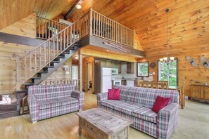 A wooden cabin interior with a staircase, two plaid sofas, a coffee table, and a kitchen area.