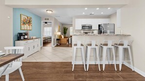 A modern kitchen with white cabinets, a central island with bar stools, and a dining area with a wooden table and white chairs. - Private Heated Pool | Lanai + Fire Pit (Largo)