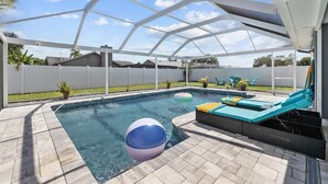 A covered outdoor swimming pool with a white roof, a lounge area with cushions, and a clear sky. - Private Heated Pool | Lanai + Fire Pit (Largo)