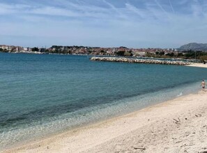 Ein Strand mit klarem Wasser, einem steinernen Wellenbrecher und einer Person, die am Sand entlanggeht.