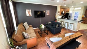Ein modernes Wohnzimmer mit einer Couch, einem Couchtisch und einem Fernseher. Es gibt eine Küchenzeile mit einer Bar und Hockern.