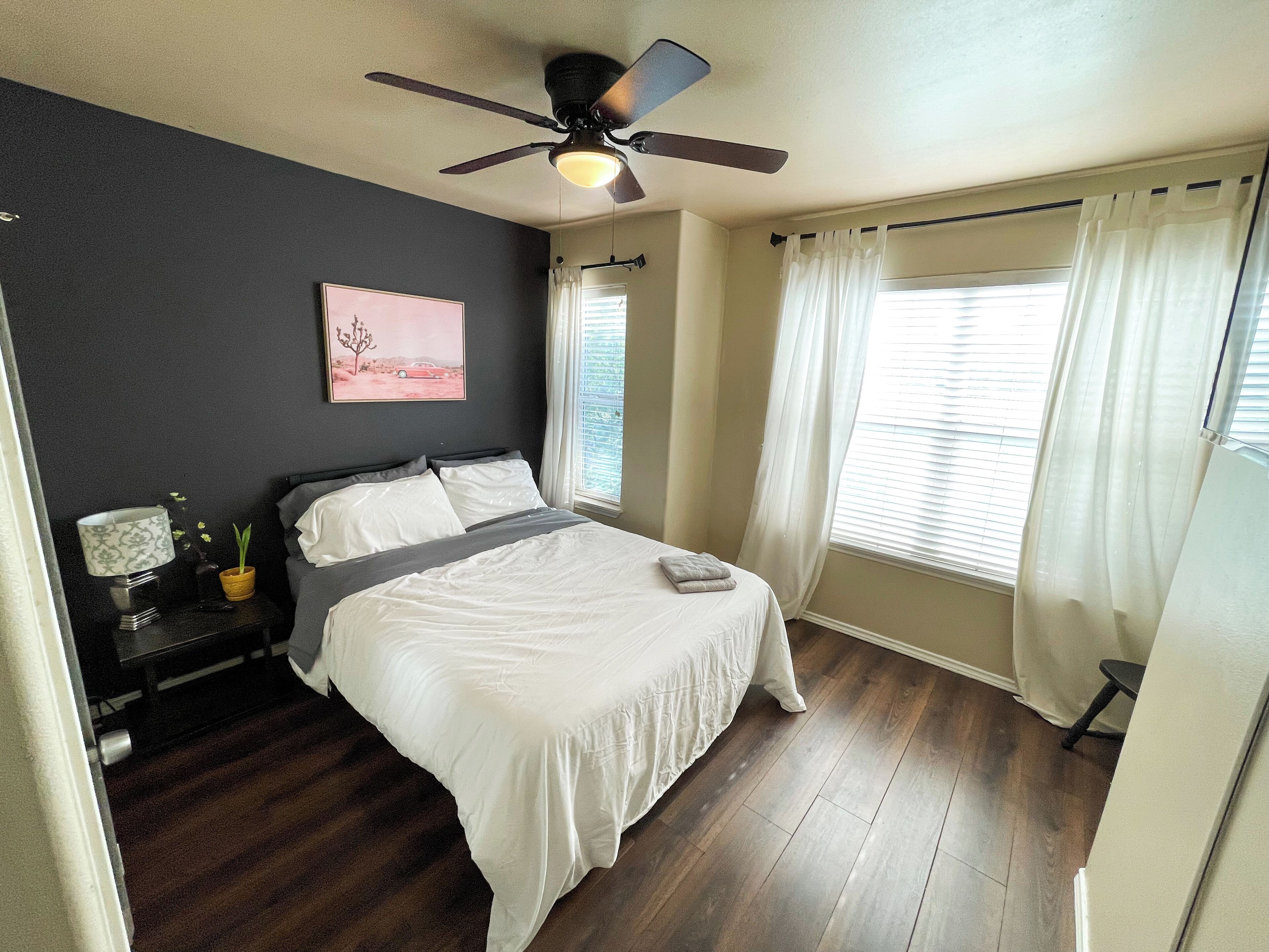 A bedroom with a bed, a ceiling fan, a nightstand with a lamp, a window with curtains, and a painting on the wall.