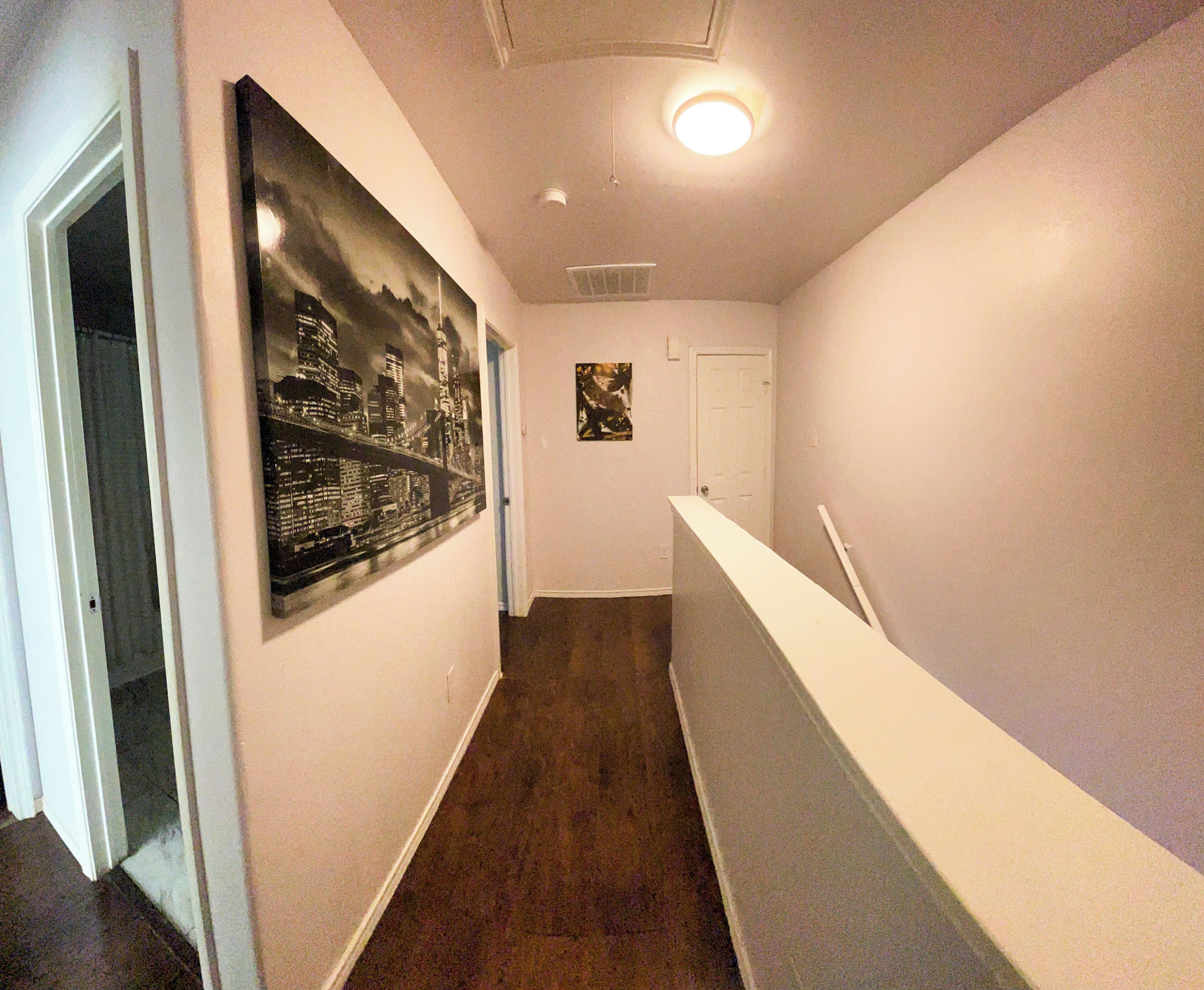 A narrow hallway with a dark wooden floor, white walls, and a white counter on the right. There is a door at the end of the hallway and a poster on the left wall.