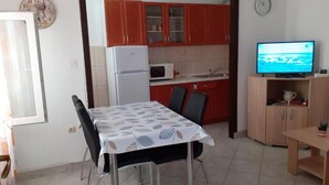 Eine kompakte Küche mit roten Schränken, einer Mikrowelle und einem Kühlschrank. Ein Esstisch mit Stühlen und ein Fernseher auf einem Holzgestell sind ebenfalls vorhanden.