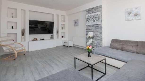 Una sala de estar moderna con un sofá gris, una silla de madera, un televisor de pantalla plana y una mesa de centro con un jarrón de flores.