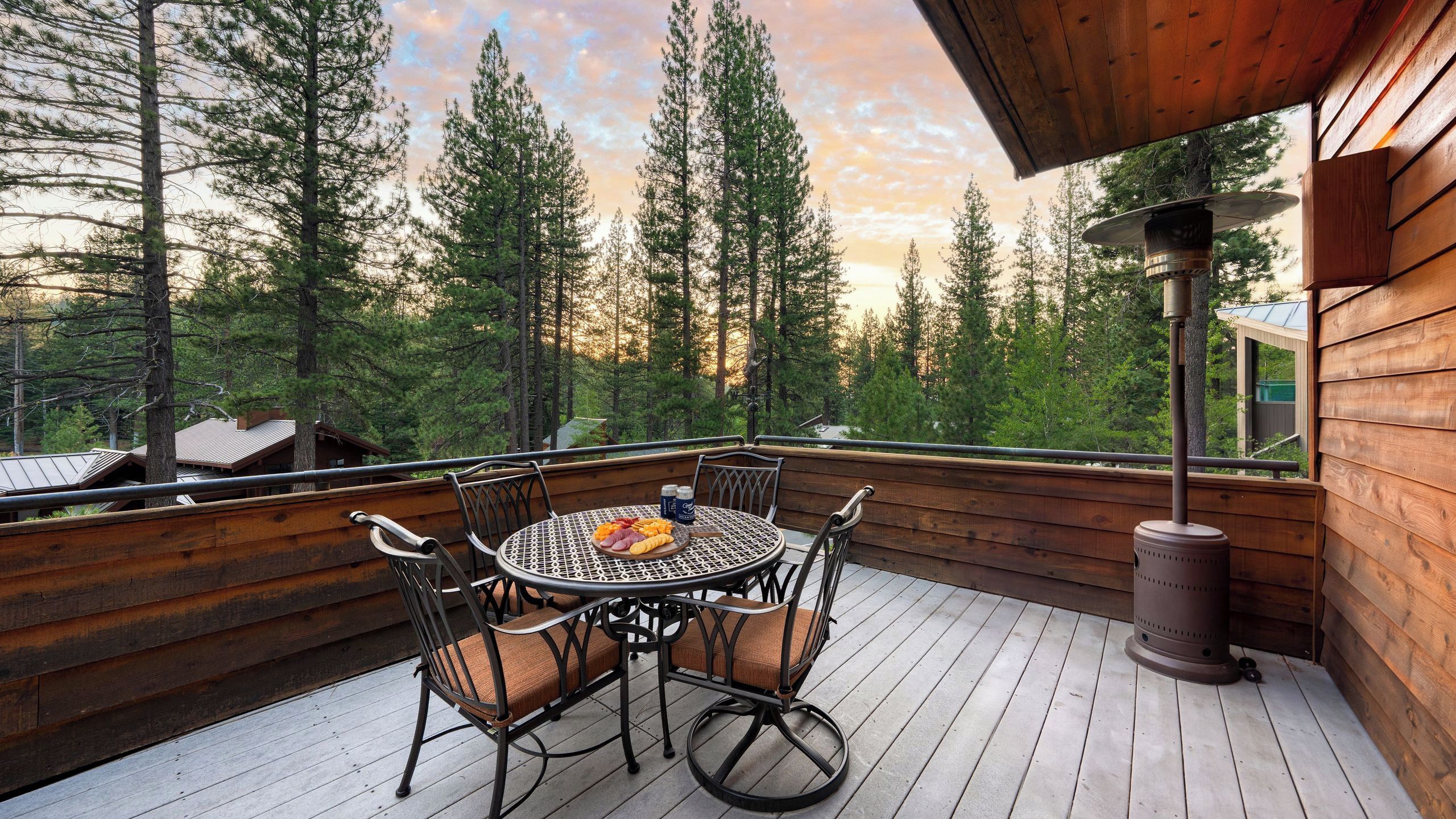 A wooden deck with a round table and chairs, surrounded by tall trees and a clear sky.