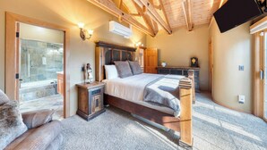 Quarto com teto de madeira, cama com cabeceira, mesa de cabeceira, cômoda, TV de tela plana na parede e uma janela grande.
