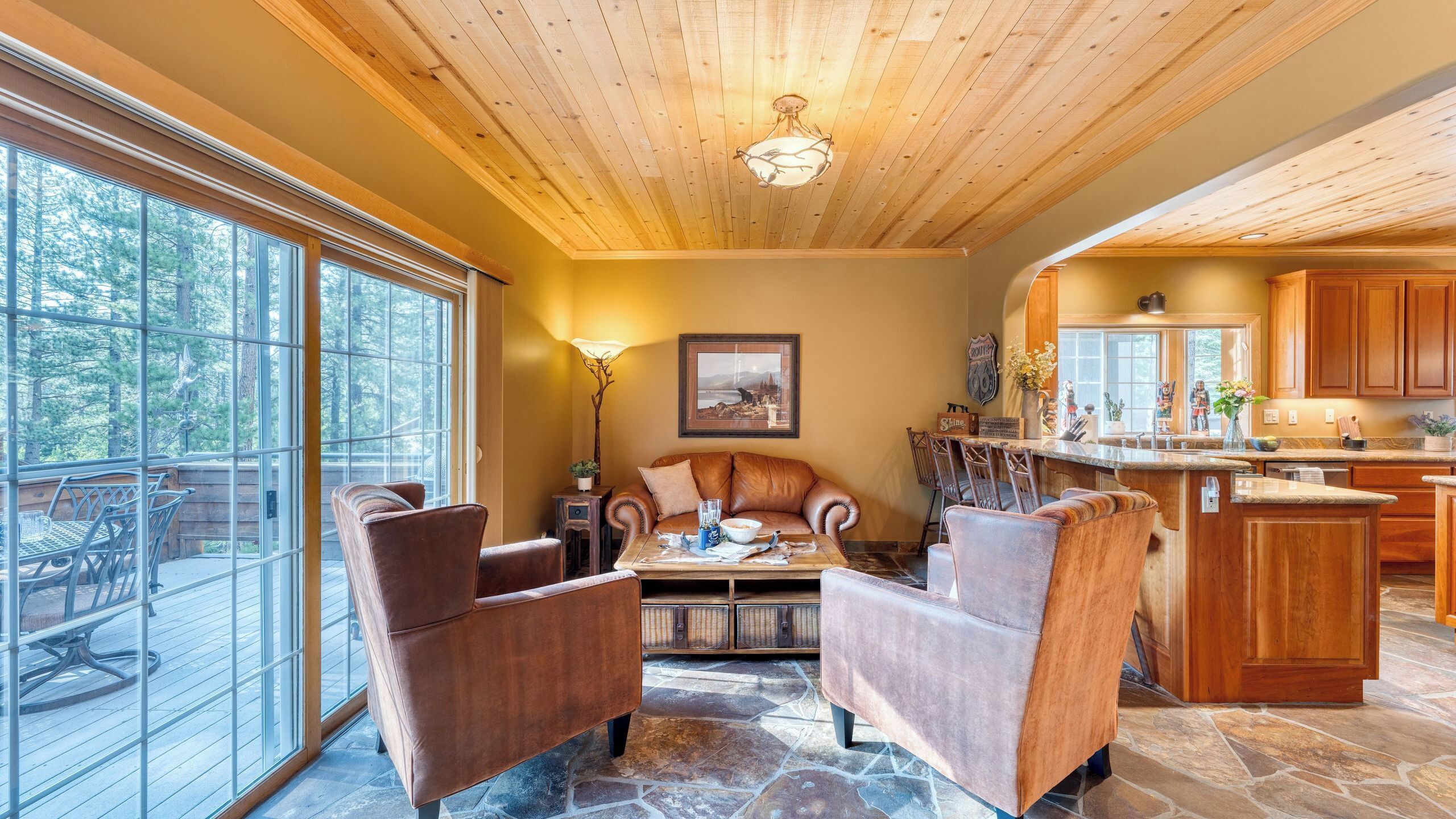 A spacious living area with a stone floor, wooden ceiling, and large windows overlooking a forest.