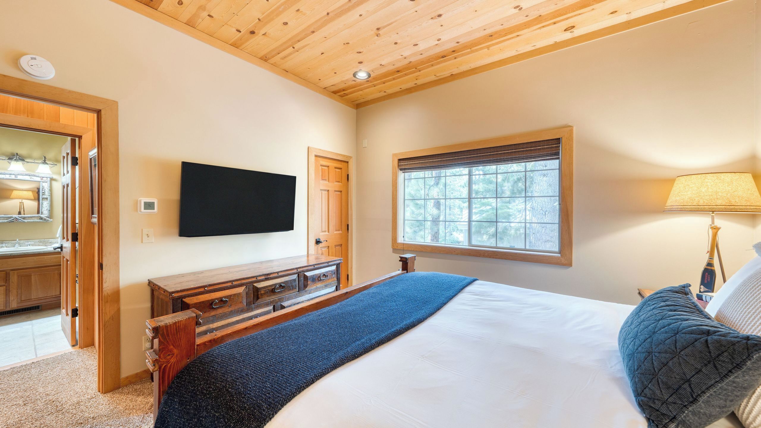 A bedroom with a bed, a TV mounted on the wall, a window with blinds, a wooden dresser, and a lamp.