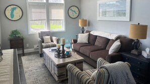 A living room with a brown sofa, striped armchairs, a coffee table, and two lamps. There are two windows with blinds, a map on the wall, and a framed picture above the sofa.
