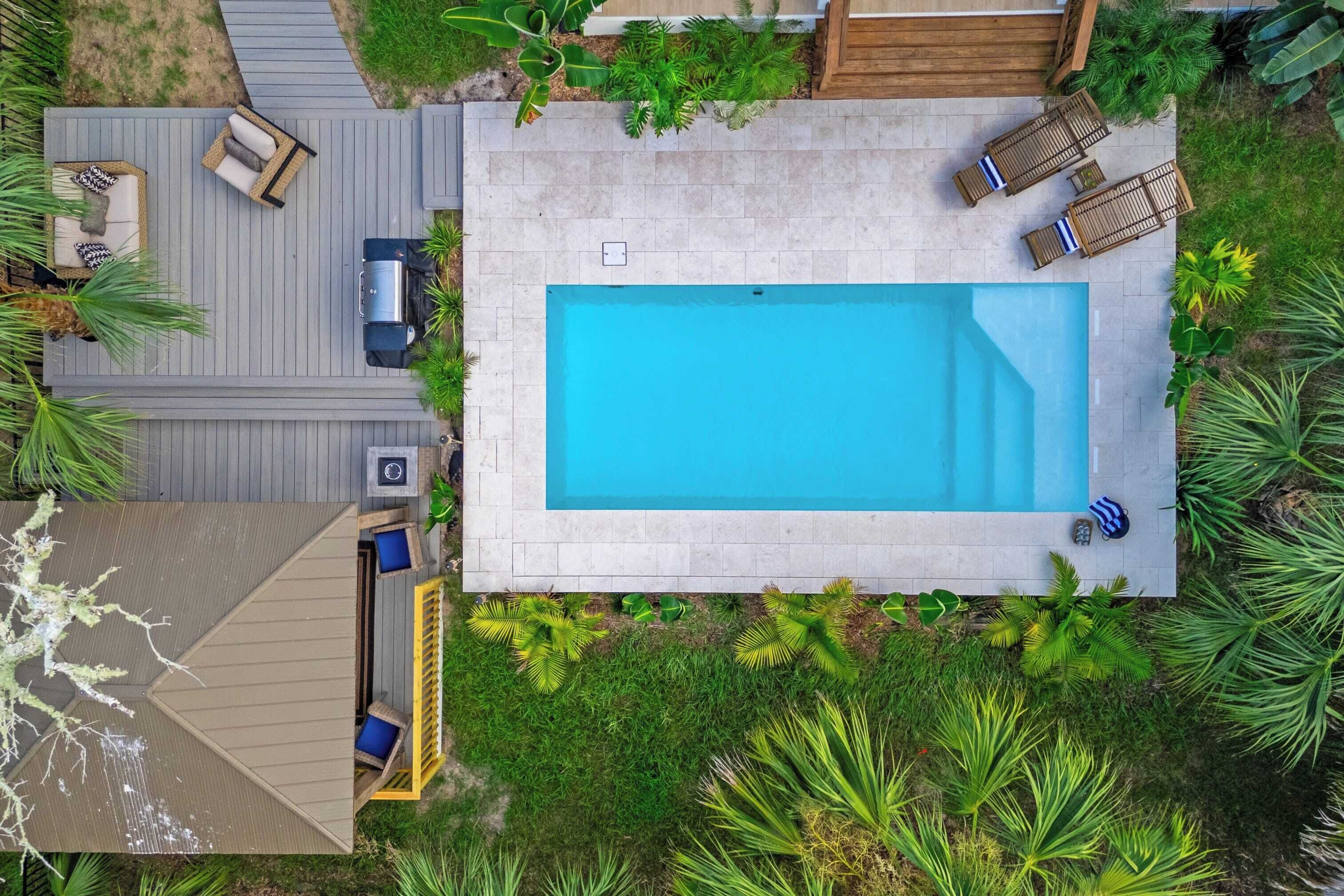 Ein rechteckiger Swimmingpool mit umlaufender Holzterrasse, ausgestattet mit Liegestühlen und einem Grillbereich.