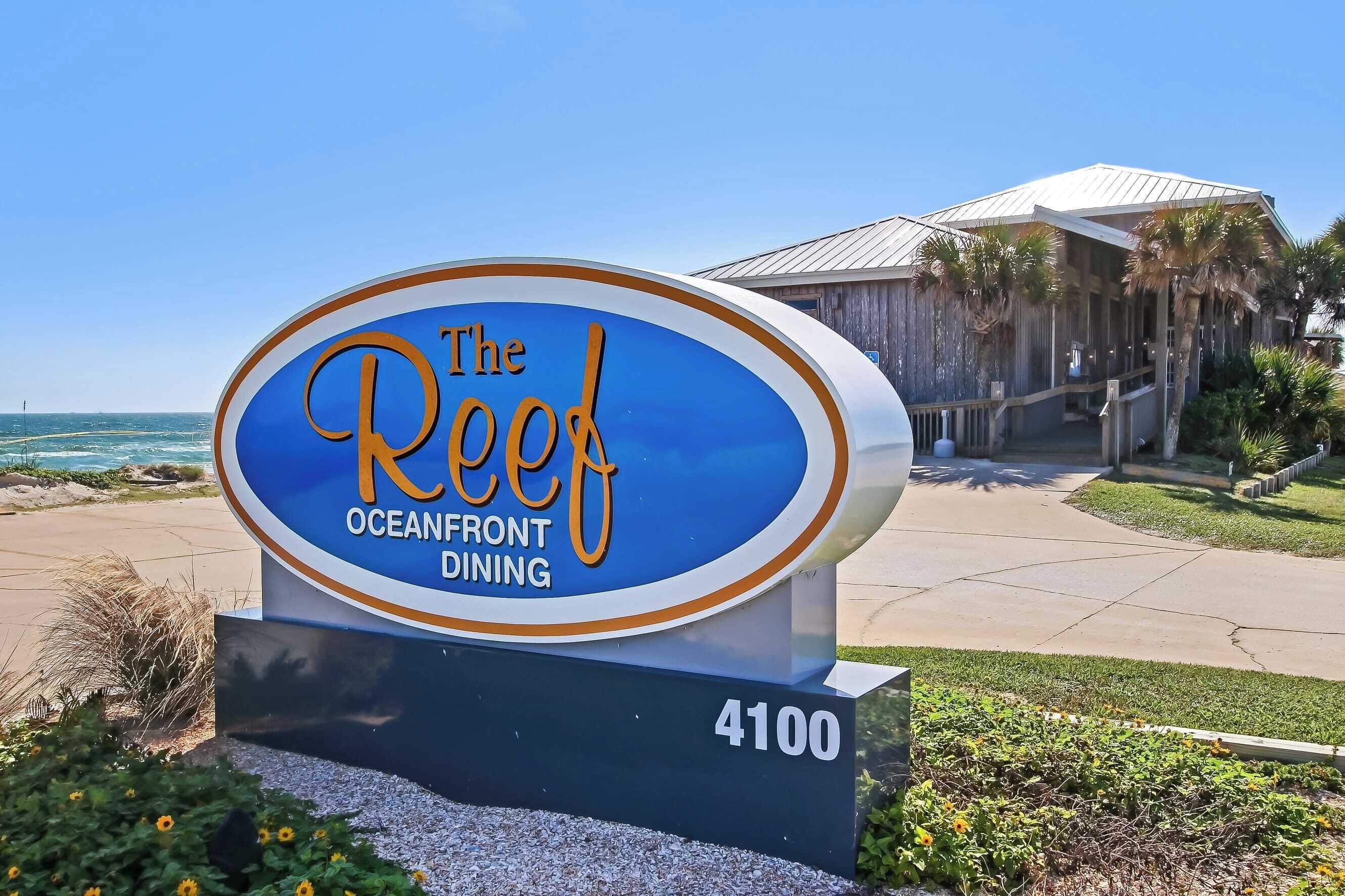 A sign for "The Reef Oceanfront Dining" with a view of the ocean and a building in the background.