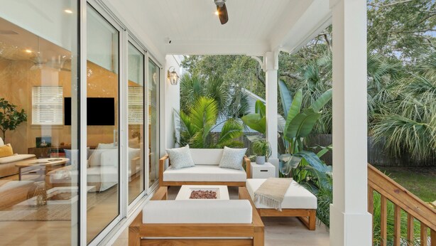 A modern living room with a glass wall, white furniture, and a view of lush greenery. - Serenity at Surfside by Avantstay Pool, Porches, Fire Pit & Arcade Near Vilano Beach (St. Augustine)