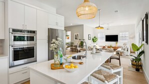 A modern kitchen with white cabinetry, stainless steel appliances, a central island with seating, and pendant lighting. - Serenity at Surfside by Avantstay Pool, Porches, Fire Pit & Arcade Near Vilano Beach (St. Augustine)