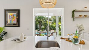 A modern kitchen with a large sink, dining area, and a view of a tropical garden. - Serenity at Surfside by Avantstay Pool, Porches, Fire Pit & Arcade Near Vilano Beach (St. Augustine)