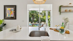 A modern kitchen with a white countertop, stainless steel sink, and a hanging light fixture. - Serenity at Surfside by Avantstay Pool, Porches, Fire Pit & Arcade Near Vilano Beach (St. Augustine)