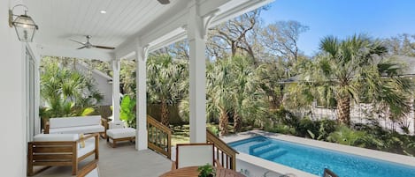 A covered outdoor area with a pool, wooden furniture, and a view of the surrounding greenery.