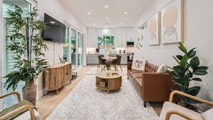 A modern living room with a sofa, coffee table, and a dining area with chairs. There is a kitchen in the background with cabinets and a TV mounted on the wall.