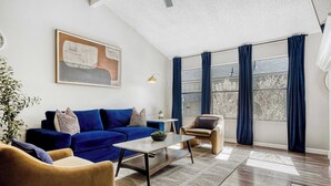 Un salon avec un canapé bleu, une table basse et un fauteuil. Il y a un tableau au mur et une fenêtre avec des rideaux.