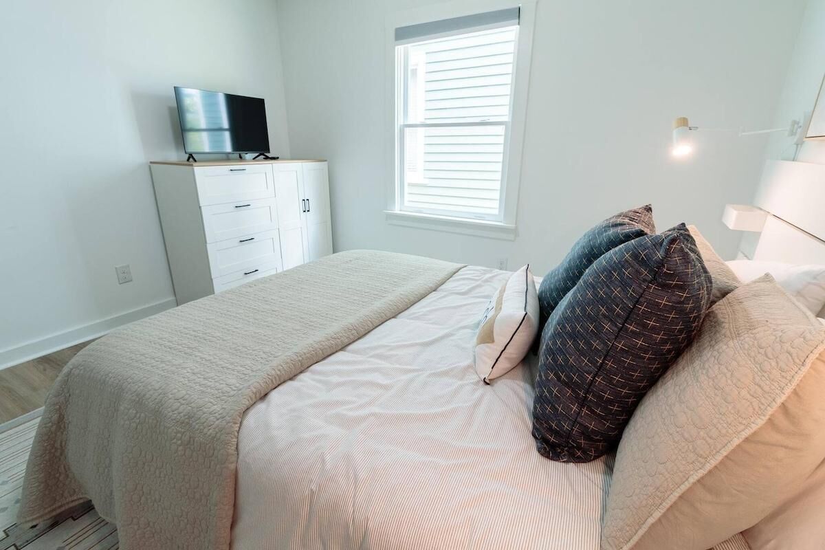 A bedroom with a bed, a TV on a stand, a window with blinds, and a bedside table with a lamp.