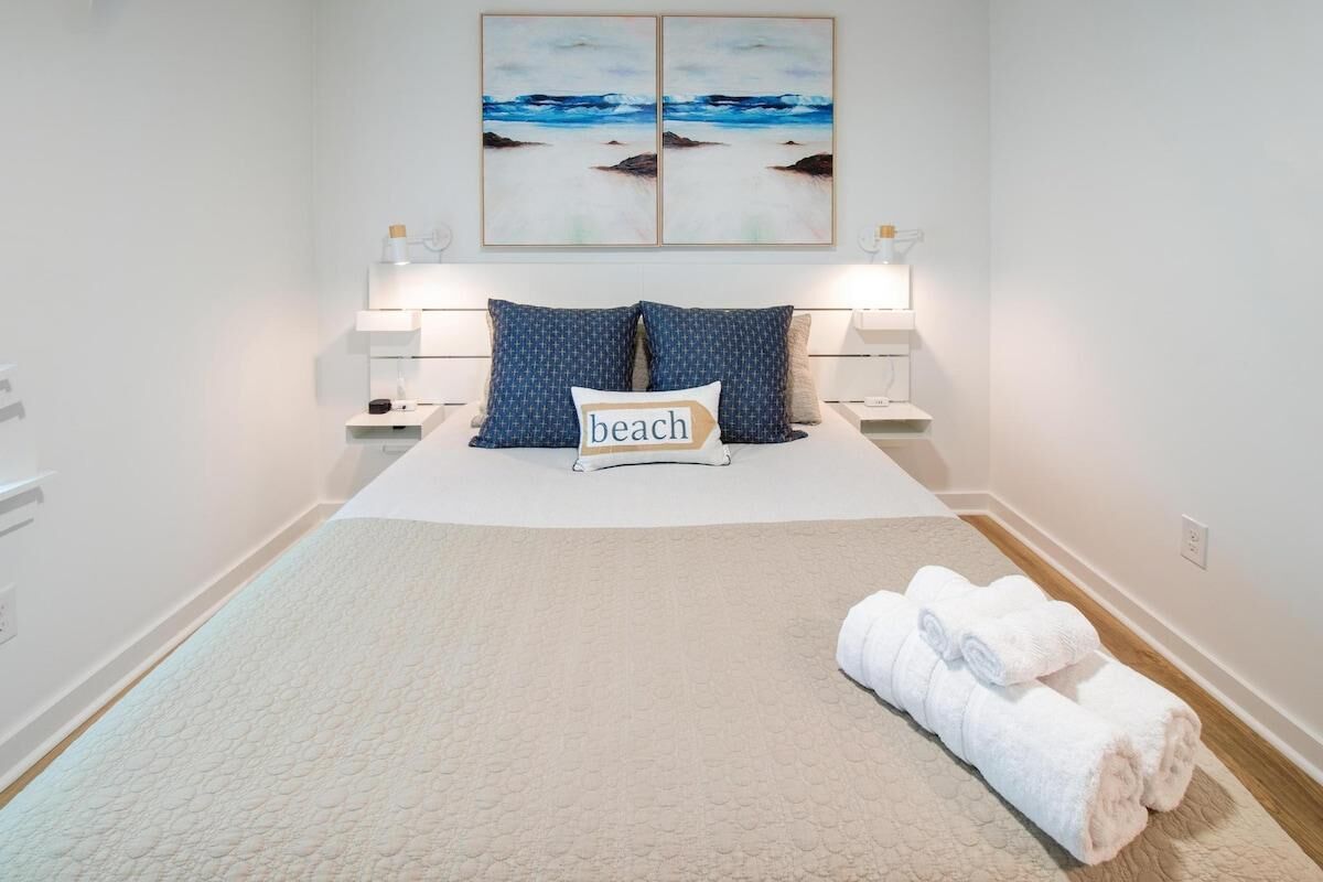 A neatly arranged bedroom with a bed, pillows, a bedside table with a lamp, and a wall-mounted artwork.