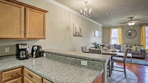 A kitchen with granite countertops, wooden cabinets, a coffee maker, and a kettle. There is a dining area with a table and chairs, and a living room with a sofa and a ceiling fan.