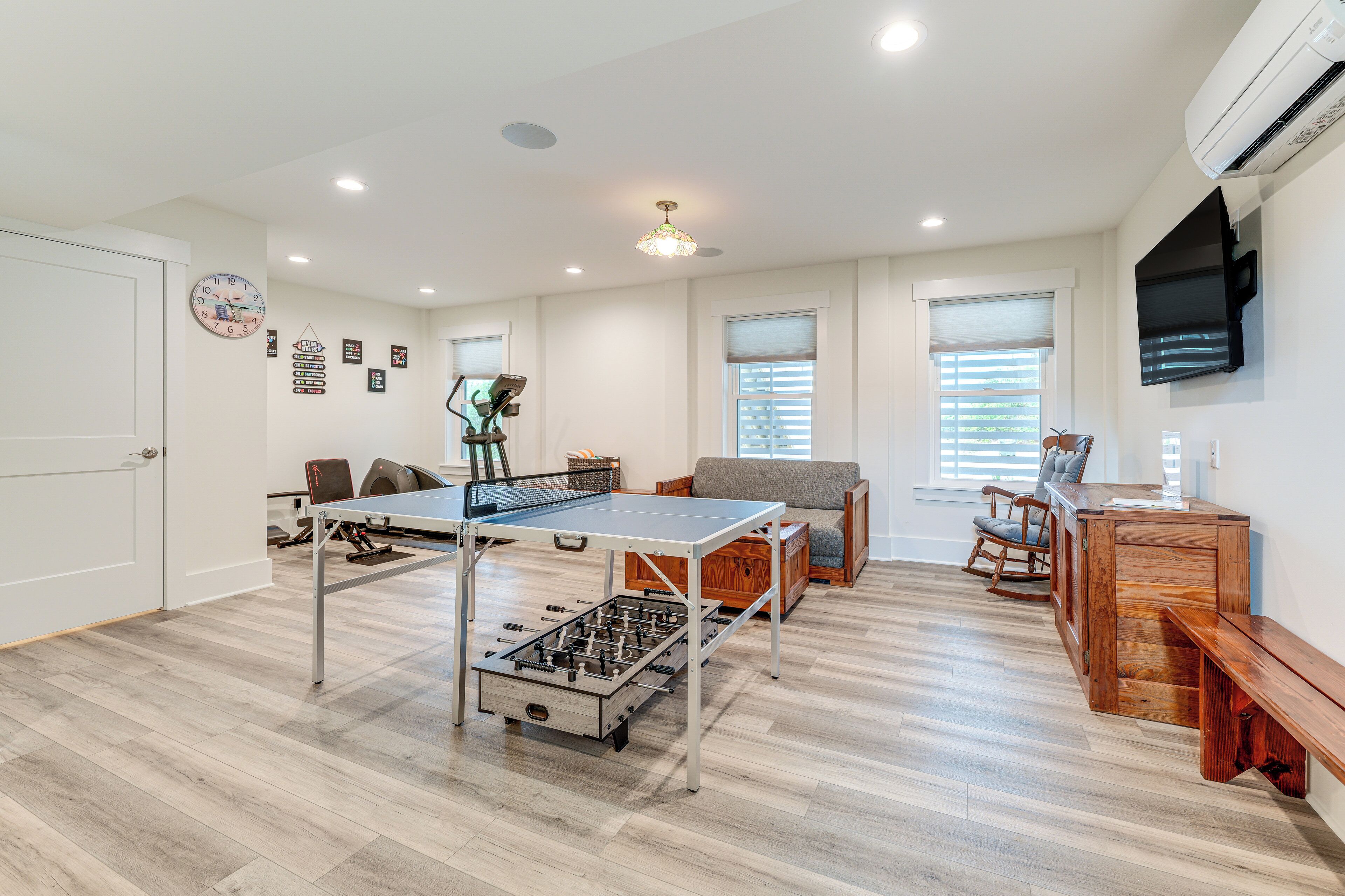 A spacious room with a ping pong table, a foosball table, a sofa, and a wooden bench. There are two windows with blinds, a television mounted on the wall, and a door.