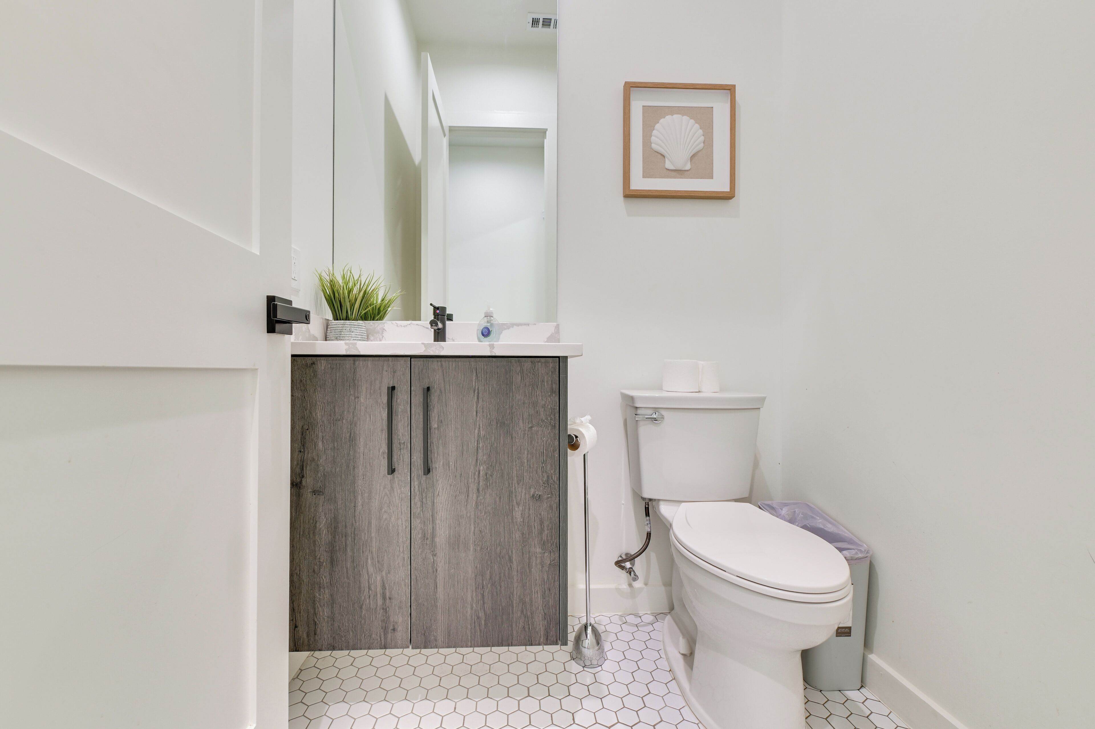 Een badkamer met een wit toilet, een houten kastje, een spiegel, een fotolijst aan de muur en een vloer van honingraatmozaïek.