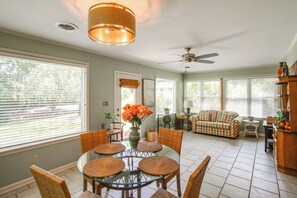 Ein Essbereich mit Glastisch und Korbstühlen, ein Wohnzimmer mit Sofa und Deckenventilator, und große Fenster mit Jalousien.