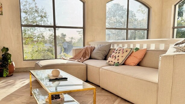 A spacious living room with a herringbone wooden floor, a glass coffee table with gold legs, a beige sofa with patterned cushions, and large arched windows offering a view of trees and a building. - Grande Casa (Stellenbosch)